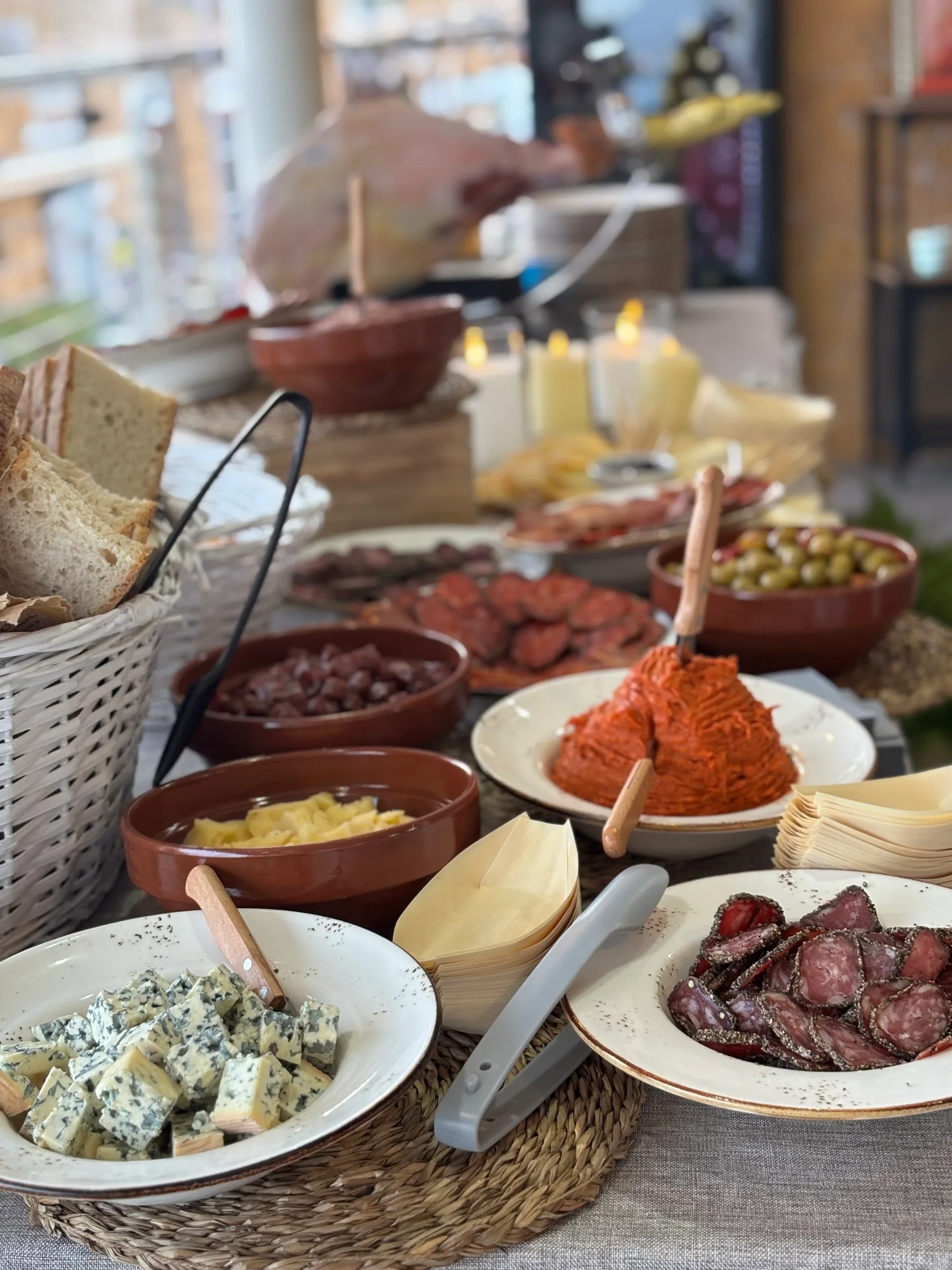 Tisch mit Käse, Salchichón, Chorizo, Oliven und Brot, dahinter wird Schinken aufgeschnitten