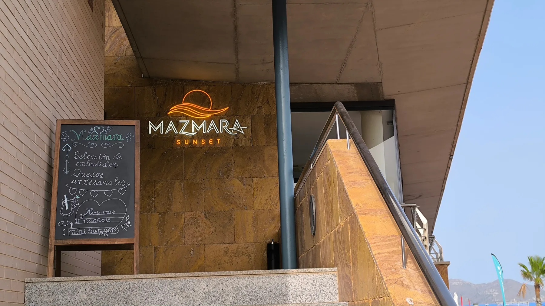 Der Eingang von Mazmara Sunset: das Neonschild mit der Sonne über der Welle und die Tafel mit der Tageskarte